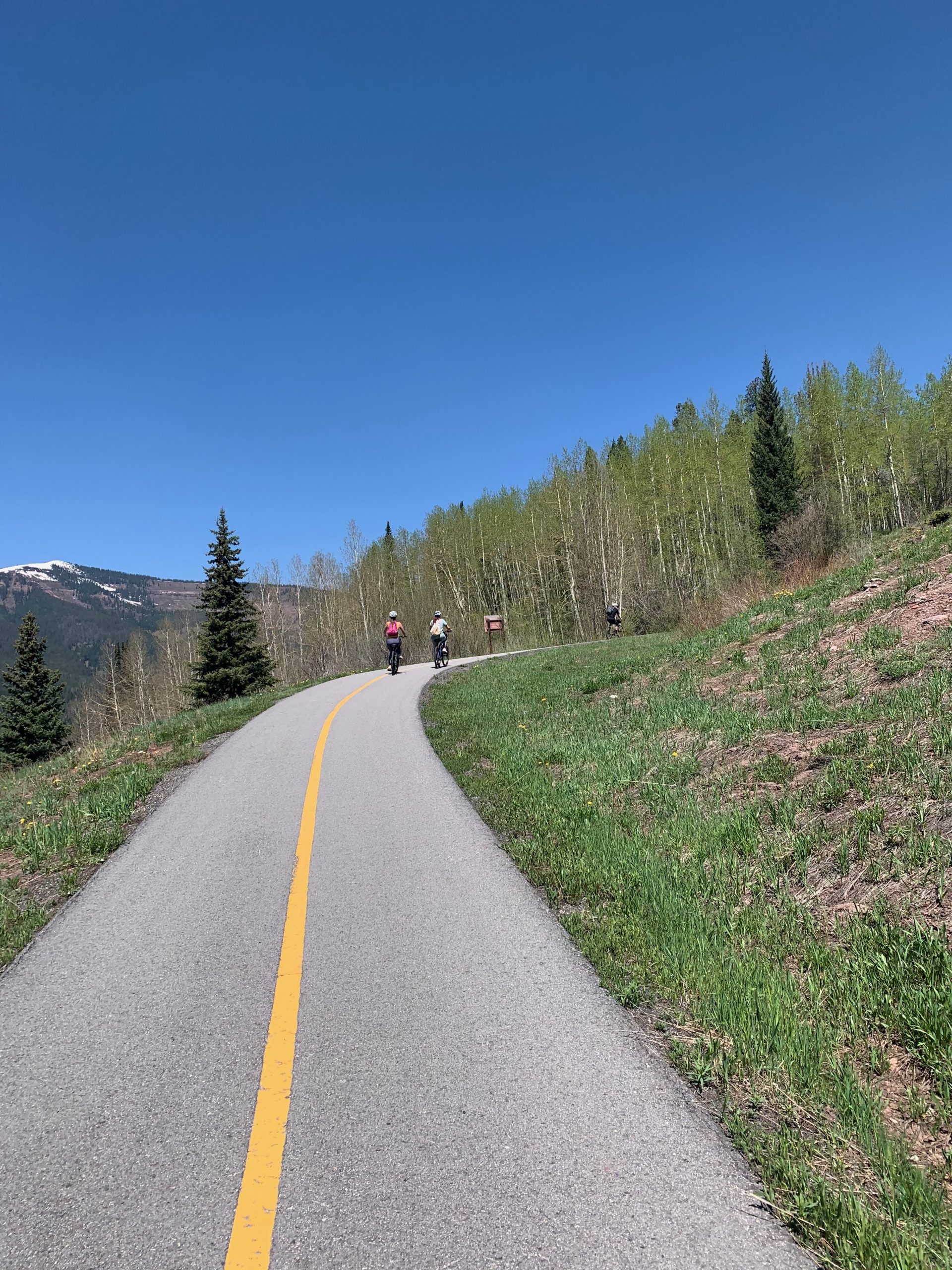 Bicyclists riding along a scenic path in Vail, Colorado, surrounded by lush greenery and blue sky, highlighting summer outdoor activities and bike rentals.