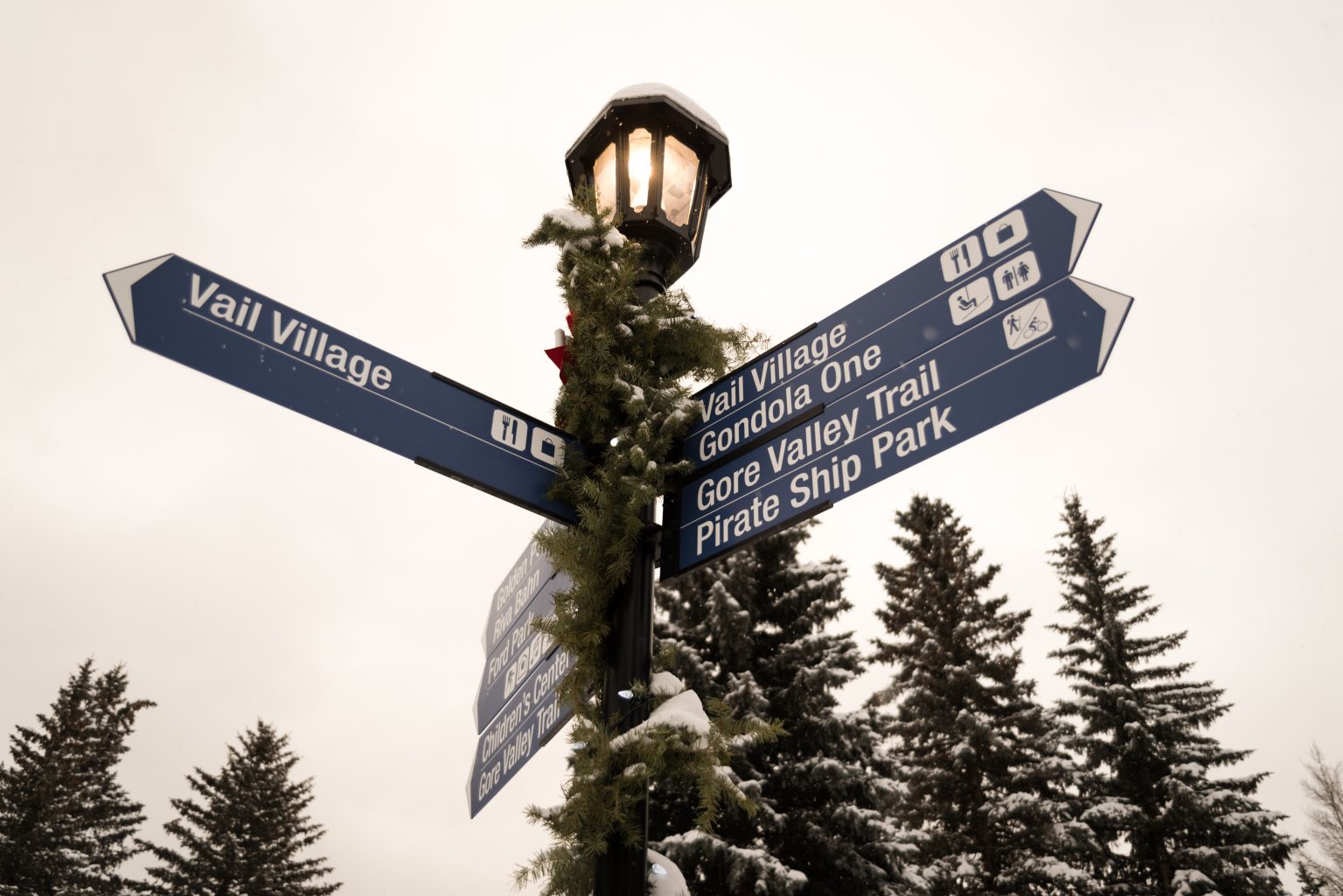 Directional signpost in Vail, Colorado, featuring signs for Vail Village, Gondola One, Gore Valley Trail, and Pirate Ship Park, adorned with holiday greenery and snow.