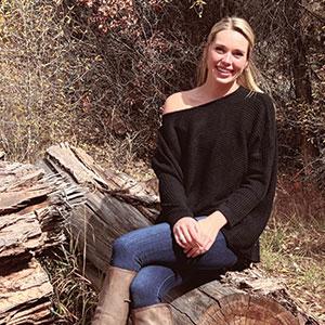 Carlee Morrison, manager at Beaver Creek Charter, sitting on a log in a natural outdoor setting, wearing a black sweater and jeans, representing the team at Charter Sports.
