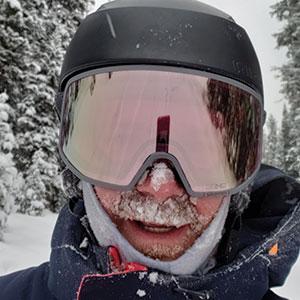 Man wearing ski goggles and a helmet, with snow on his mustache and beard, in a snowy mountain environment, representing outdoor winter sports and skiing expertise at Charter Sports.