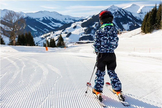 Child skiing on groomed snow slope, wearing colorful ski gear, with snow-capped mountains in the background.