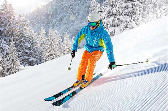 Skier in bright blue and orange gear skiing down a snowy slope surrounded by pine trees.