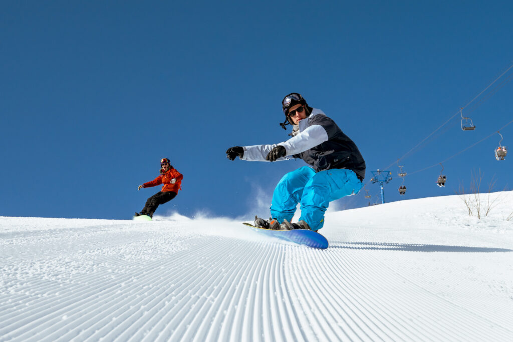 Two snowboarders carving down a snowy slope under a clear blue sky, showcasing the excitement of winter sports, relevant to seasonal job opportunities at Charter Sports.