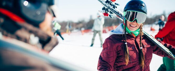 Smiling skier wearing helmet and goggles, holding skis, with snowy background and other skiers at Beaver Creek Resort.
