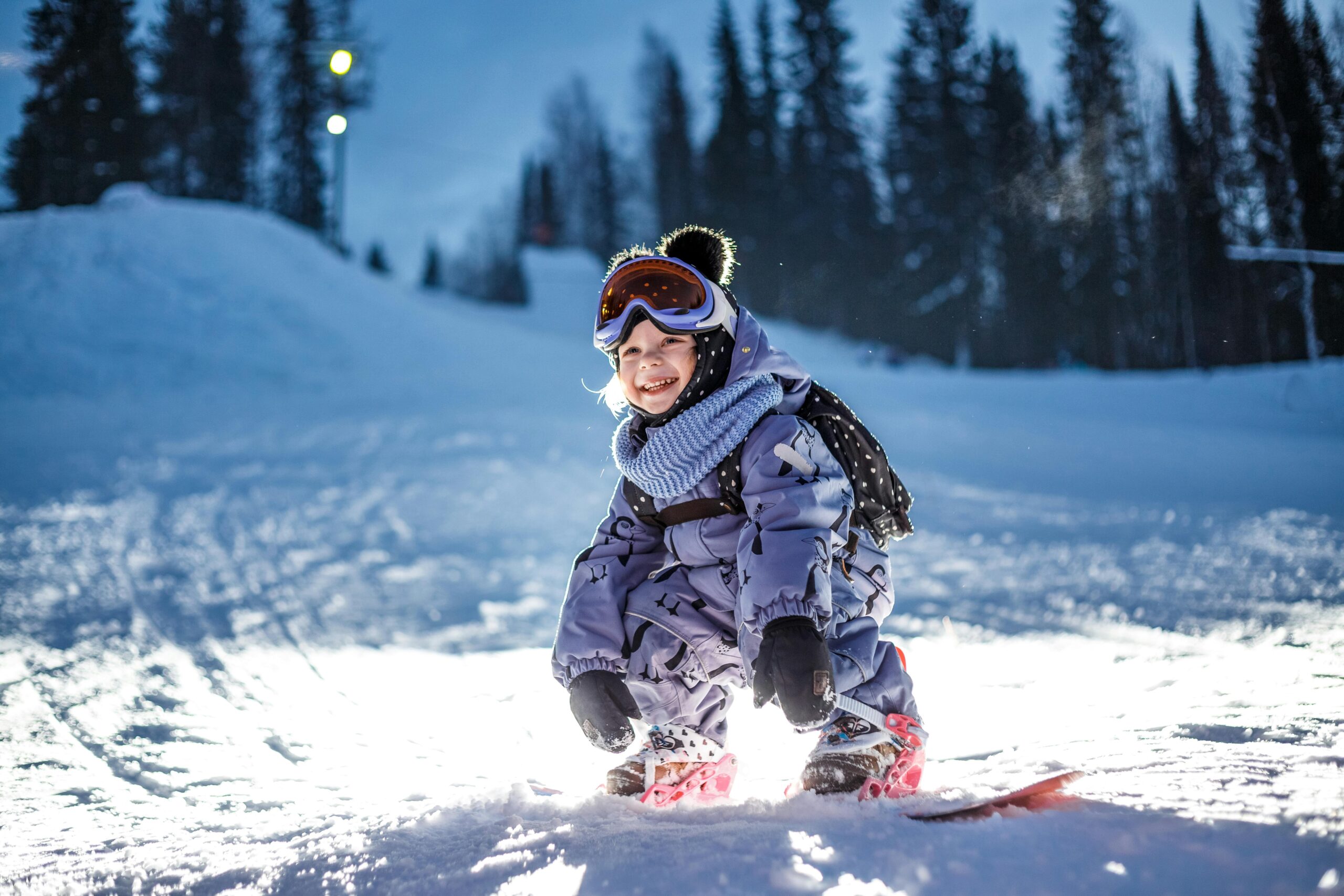 Child in winter gear smiling while snowboarding on a snowy slope, showcasing a fun outdoor skiing experience at a ski resort.