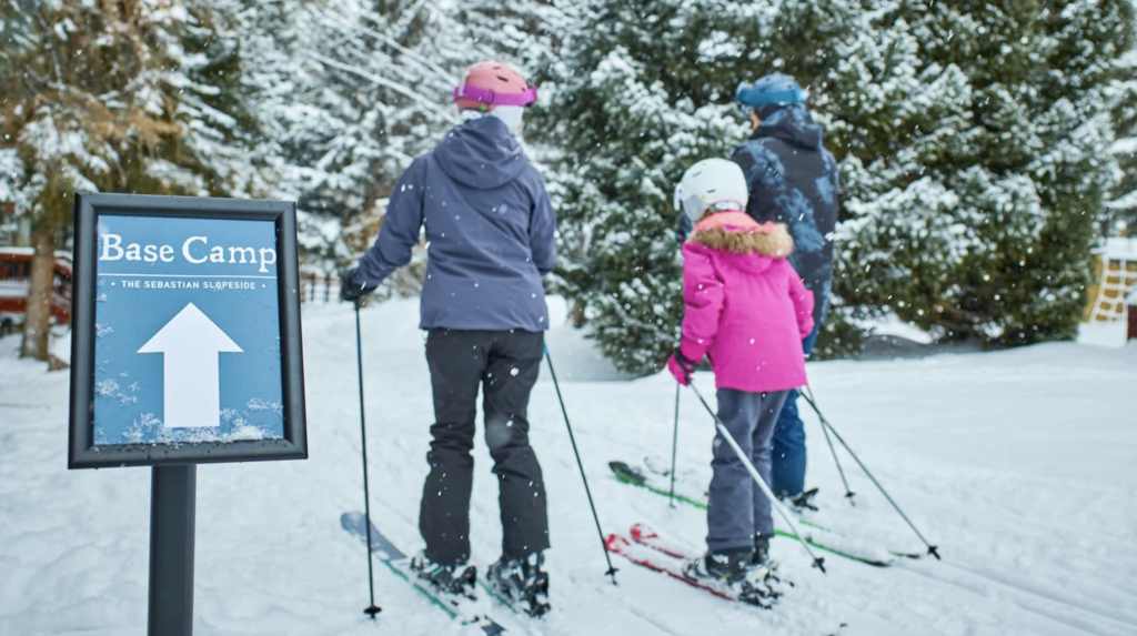 Skiers at Base Camp sign, preparing for slopeside skiing in snowy environment, emphasizing convenience of direct access to ski rentals.